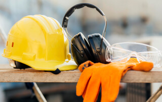 Capacete de segurança amarelo, protetor auricular preto, óculos de proteção transparentes e luvas de proteção laranja sobre uma superfície de madeira, representando o uso obrigatório de EPI no ambiente de trabalho.