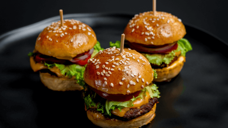 Close-up de três mini hamburgueres com pão de gergelim e queijo, servidos em um prato preto, ilustrando um rodízio de hambúrguer.