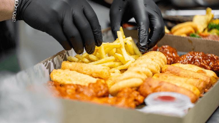 Close-up de mãos com luvas pretas arrumando uma caixa de rodízio com petiscos variados, incluindo batata frita, sticks de queijo empanados e coxinhas de frango com molho.