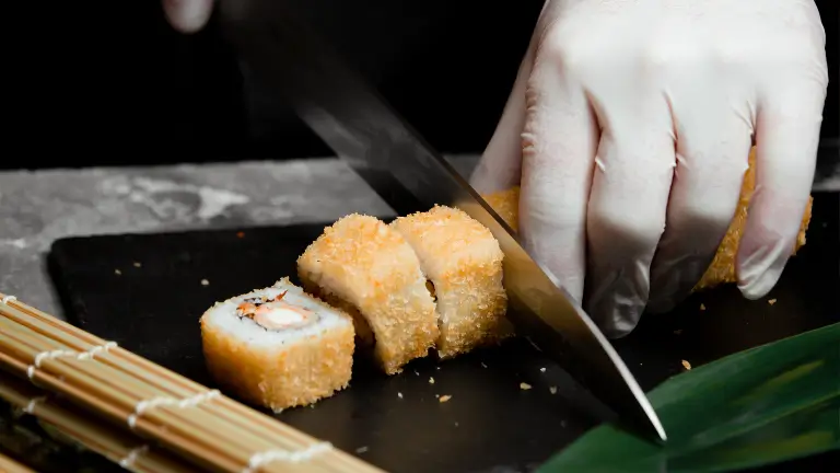 Mãos de um sushiman em um rodízio japonês cortando um rolo de hot roll (sushi frito) com uma faca profissional em uma esteira de bambu.