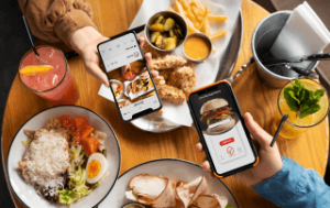 Pessoas utilizando smartphones para fotografar pratos de comida e hambúrguer em uma mesa, ilustrando o marketing para restaurantes.