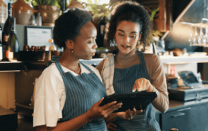Duas mulheres em um restaurante usando aventais listrados e analisando dados em um tablet digital em um ambiente de balcão, IA para restaurantes.