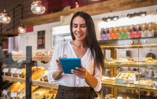 Mulher sorridente utilizando um tablet com sistema para padarias, com vitrines de pães e doces ao fundo.